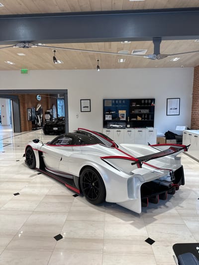 The image displays a high-performance Pagani hypercar within a luxurious showroom environment, likely located in Greenwich, United States. The central subject is a pristine white Pagani, possibly a Huayra or Zonda model, distinguished by its intricate red and black aerodynamic detailing. The car's unique gullwing doors are open, revealing a glimpse of its dark interior. It rests on a polished, light-colored marble tiled floor, which includes sporadic dark square inlays. To the left, the flooring transitions to light-toned wooden planks.
The setting is a sophisticated dealership or showroom with clean white walls. Two framed monochrome artworks depicting car designs are visible on the wall. Behind the car, a display cabinet showcases various luxury items, including what appears to be a bottle of champagne, car models, and other merchandise. The brand name "PAGANI" is clearly visible on one of these displays, along with the partially discernible word "TRAGON" or "RAGON." A prominent dark grey structural beam with attached metal tension cables runs vertically on the left side of the frame. The area is well-lit by recessed ceiling lights and subtle wall-mounted fixtures.
In the background, a large doorway opens to another section of the facility, where more vehicles are faintly visible. A green "EXIT" sign is present above this doorway. The scene conveys a sense of luxury and exclusivity, with the car presented as a centerpiece of automotive artistry. There are no people visible in the image.