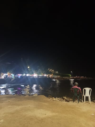 The image shows a nighttime scene at the waterfront in Tolú, Colombia. In the foreground, a sandy area with scattered rocks leads to the water. Two plastic chairs are placed on the sand, with one occupied by a person wearing a pink and black shirt and a baseball cap, facing the water. To their right is an empty white plastic chair. The water's surface reflects the lights from the city. In the background, a line of buildings and stalls is illuminated by various colorful lights, indicating a commercial area along the waterfront. Palm trees are visible amongst the buildings. Further in the distance, streetlights and more lights from the city are scattered along the coastline. The sky is dark, suggesting it is late evening or night.