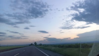 The image shows a view from inside a vehicle, looking out through the windshield at a road stretching into the distance. The road appears to be a paved highway with lane markings. On either side of the road, there is green vegetation, likely fields or meadows. In the distance, there is a line of trees and possibly a low mountain range. The sky is filled with clouds, suggesting twilight or dusk. The image has some smudges and imperfections on the windshield, obscuring the view slightly. The image was taken near Sokolovo, Bulgaria.