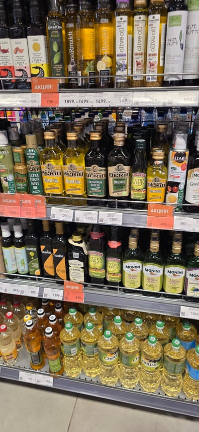 This is a vertical, eye-level, medium shot of a well-stocked supermarket shelf filled with various types of cooking oil. The scene appears to be indoors, likely in a grocery store, as indicated by the organized rows of products and shelf pricing. The lighting is bright and even, typical of a commercial setting.
The main subjects are numerous bottles of cooking oil, predominantly olive oil and sunflower oil. The bottles vary in size, shape, and color, with labels displaying brand names and product types in different languages, including Russian and English. Some bottles are clear, revealing the golden liquid inside, while others are dark green glass.
Several bottles are marked with bright orange "АКЦИЯ!" (meaning "SALE!" or "PROMOTION!") tags, indicating special offers. Some of the visible brands include "Filippo Berio," "MONINI," "BORGES," "Casa Rinaldi," "Costa d'Oro," and "TERRA DELVESSA." The sunflower oil bottles on the right side of the frame are larger and have green caps, while many of the olive oil bottles have gold or black caps.
The setting is a retail environment, likely a food section of a supermarket. The shelving is metallic and appears to be well-maintained. The overall impression is one of abundance and a wide selection of products for consumers. No people are visible in the image, and there are no cues about the time of day or weather. The dominant emotion evoked is one of everyday shopping and consumer choice.
Visible text includes brand names, product descriptions like "EXTRA VIRGIN OLIVE OIL," "ORGANIC OLIVE OIL," "OLIVE OIL," and "SUNFLOWER OIL," as well as pricing information, such as "739,00," "1399,00," "1049," and "559." The "АКЦИЯ!" labels are also prominent text.
