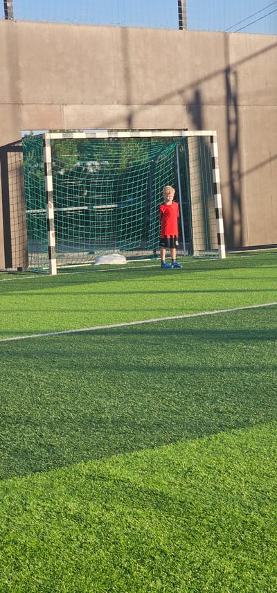 A young boy stands on a green artificial turf soccer field in front of a soccer goal. The boy is wearing a red t-shirt and black shorts. He has blond hair and is facing the camera. The soccer goal has a green net and a white frame. In the background, there is a gray wall and a blue sky. The scene is lit by sunlight, casting shadows on the field and wall. The location is the city of Овощной, Russia.