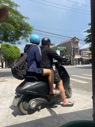 Two individuals are seated on a black electric scooter, traveling on a paved road. The person in front is wearing a blue helmet and a dark blue hoodie with white stripes on the sleeves, carrying a black backpack. The person behind is wearing a black helmet and a black long-sleeved shirt, with black shorts. Both are wearing sandals. The scooter has a license plate that reads "26.75 A01 426.75". The environment appears to be a street scene in Tân Quang Commune, Vietnam, with buildings, trees, power lines, and a crosswalk visible. The sky is partly cloudy.