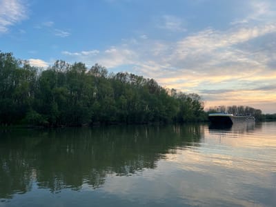 The image depicts a river landscape in Harsova, Romania, at dusk or dawn. A large, flat-topped barge is on the river, parallel to the shore. The shore is lined with trees exhibiting spring foliage. The water surface is reflective, mirroring the sky and shoreline. The sky exhibits a blend of blues and yellows. No people are visible.