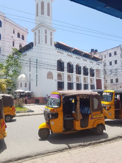 The image depicts a daytime urban street scene in Majengo ward, Kenya, under a clear blue sky. Dominating the background is a prominent white multi-story building featuring a distinct minaret with patterned windows, indicative of a mosque or Islamic cultural center. This building displays arched windows on its lower levels, rectangular windows with dark shutters on upper floors, and a rooftop balcony area. To the left, a section of another white building is visible, and to the right, a less ornate, lighter-colored multi-story building with multiple dark rectangular windows.

On the asphalt road in the foreground and middle ground are three yellow three-wheeled auto-rickshaws, commonly known as tuk-tuks. The central tuk-tuk, with a brown canopy, is positioned facing slightly left, displaying "TAXI" text on its side and purple trim on its windshield. Its passenger compartment is visible, containing a striped fabric item. A second yellow tuk-tuk is partially visible on the far left. A third yellow tuk-tuk is parked or stationary further to the right, with its passenger door open.

Several indistinct figures are discernible on the sidewalk near the base of the prominent white building, some positioned near small white umbrellas, suggesting vendors or pedestrians. Green foliage from a tree or bush is visible near the left side of the main building. Overhead, multiple black power lines traverse the sky. The scene is brightly lit by sunlight, casting shadows.