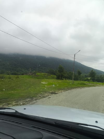 The image depicts a landscape scene from inside a vehicle, looking out towards a mountain range under a cloudy sky. In the foreground, the dashboard and windshield wipers of the car are visible. The road curves to the right, with a gray, asphalt surface. To the left of the road, a grassy hill rises, dotted with some rocks. In the mid-ground, a wooden cross monument stands on the hill, surrounded by a low stone wall. Further back, a dense forest covers the lower slopes of the mountain. The upper portion of the mountain is shrouded in thick, gray clouds, obscuring its peak. A single lamppost stands near the road on the right side of the image. The overall atmosphere is overcast and somewhat somber. The image appears to be taken in the vicinity of Skurdi, Georgia, given the characteristic mountainous terrain and lush vegetation.