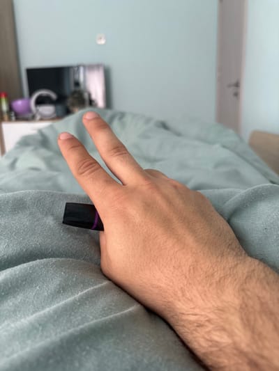 A person's right hand is shown making a "peace" or "victory" sign while holding a small, black and purple object (likely a tube of lipstick or similar cosmetic product) between the ring and middle fingers. The hand is resting on a light blue/grey fabric that appears to be clothing or bedding. The background is blurred, showing a light blue wall, a piece of furniture, and a small object. The setting doesn't appear to be Nadarevo, Bulgaria.