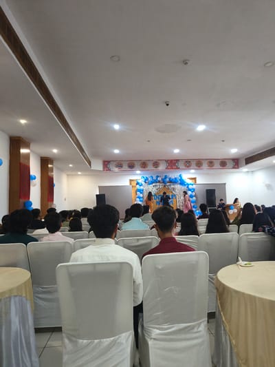 The image depicts an indoor event, possibly a graduation or award ceremony, taking place in a venue in Khaniara, India. The room is filled with an audience seated in rows of white-covered chairs, facing a stage area. The stage is decorated with a balloon arch in blue and white, and a metallic fringe curtain. Several people, including performers and organizers, are visible on the stage. The ceiling features recessed lighting and ventilation. The overall atmosphere suggests a formal or semi-formal gathering.