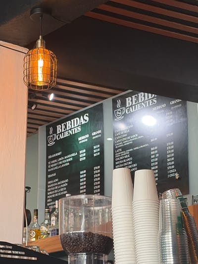 The image showcases cafe menus featuring "Bebidas Calientes" (Hot Drinks). One menu lists coffee options like Americano, Café de Olla, and Cappuccino, along with Lattes (caramel, vanilla, amaretto, Golden Milk), and Mochas. Another menu features gourmet teas, including green tea with lemon and chamomile, black tea with red fruits, and chamomile with lavender. Additionally, there are "Tisanas" (herbal teas) like strawberry kiwi, apple cranberry, passion fruit, blackberry, and guava punch, with prices listed for "Mediano" (medium) and "Grande" (large) sizes.