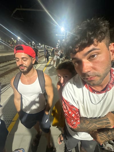 The image depicts a group of people on a train platform at night, likely in El Masnou, Spain, given the context of the provided crops which hint at Spanish signage. The central focus is a man in the foreground, wearing a white tank top, dark shorts, and a red baseball cap. He has a backpack on and is wearing a necklace with a red pendant. To his left, another man with a red cap and white tank top is visible, looking down. Behind the man in the foreground, a woman with light brown hair is peeking from behind him, looking directly at the camera. In the background, other people are standing on the platform, some with their backs to the camera, and train tracks are visible. The platform is illuminated by artificial lights, creating strong highlights and shadows. The overall scene suggests a casual gathering or travel at a public transportation hub during nighttime hours.