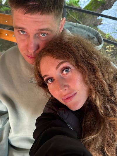 A selfie-style photograph depicts a fair-skinned couple with light eyes. The woman, positioned on the upper right, has shoulder-length curly auburn hair and is wearing a black jacket. The man, on the lower left, has short brown hair and is wearing a light green hooded sweatshirt. The location appears to be outdoors with trees and a possible structure visible in the upper left background. The image lacks the necessary details to definitively confirm the location is in Abzhanda, Abkhazia.