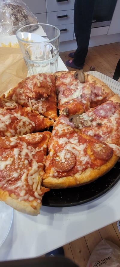 A close-up, overhead shot shows a partially eaten pizza on a black circular plate. The pizza is cut into several slices and features toppings such as pepperoni, mushrooms, ham, and melted mozzarella cheese. To the left of the pizza is a clear glass filled with a clear liquid, possibly water, resting on a brown paper bag. In the background, a person wearing black pants and purple slippers stands near kitchen cabinets and a silver appliance. The scene appears to be in a domestic kitchen setting, possibly in Dietzenbach, Germany, as indicated by the text on a bag in the lower right corner which includes "Ganz op". The lighting suggests an indoor environment, likely daytime.