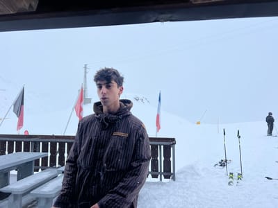 The image depicts an outdoor, snow-covered scene identified as Valtournenche, Italy, under active snowfall. A young male, positioned slightly left of center, stands facing the camera. He has dark, wavy hair and wears a dark brown, vertically striped hooded jacket with a rectangular patch containing text on its upper right chest area. Snowflakes are visible on his hair and jacket.
To the subject's left, a wooden picnic table with benches is partially covered in snow. A dark wooden railing forms a barrier behind the subject and the table. Beyond the railing, three flags are visible: a black and red flag on the left, a red and white flag to its right, and a French national flag (blue, white, red vertical stripes) further right. A metal lattice transmission tower or ski lift pylon stands in the mid-ground, left of center.
On the right side of the frame, two sets of skis with poles are upright in the snow. Further into the background, another individual in dark ski attire is visible, standing on skis and facing away from the viewer on a snow-covered slope. The entire background consists of snow-covered mountainous terrain under an overcast, light gray sky, with numerous snowflakes visible throughout the air. Yellow and red ski markers are sparsely distributed on the distant slope.
