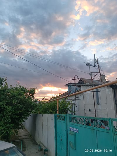 The image depicts a street scene in Изумрудновское сельское поселение, Ukraine. The sky is filled with dramatic clouds, tinged with the colors of sunset or sunrise, ranging from pale pinks and oranges to grays. Power lines crisscross the upper portion of the frame. In the middle ground, a two-story building with a damaged roof and various antennas and satellite dishes is visible. A light-colored fence runs along the left side of the image, partially obscured by dense green foliage. A turquoise metal gate, adorned with decorative scrollwork, dominates the right side of the foreground. A street sign on the gate reads "улица ЦВЕТОЧНАЯ 16" (Street Tsvetochnaya 16). The date and time "20.06.2026 20:16" are superimposed on the bottom right corner of the image. A simple wooden bench is positioned near the fence. A portion of a car is visible in the lower left corner. The overall impression is of a residential area during twilight.