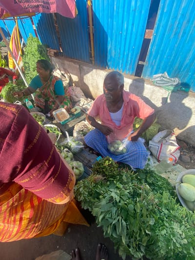 At a market stall in Chinnambedu, India, two individuals are engaged in selling produce. A woman in a teal blouse and a patterned sari sits behind a display of green vegetables. To her right, a man in a pink shirt and a blue checkered lungi is packaging some green fruits, possibly guavas or small gourds, into a plastic bag. He is seated on the ground, surrounded by sacks and more produce. In the foreground, the back of another person, dressed in a maroon and yellow striped garment, is visible. The stall is covered by a pink and yellow striped awning, with a blue corrugated metal wall providing a backdrop. The ground is paved with concrete and dirt. The scene appears to be illuminated by natural sunlight, casting shadows.