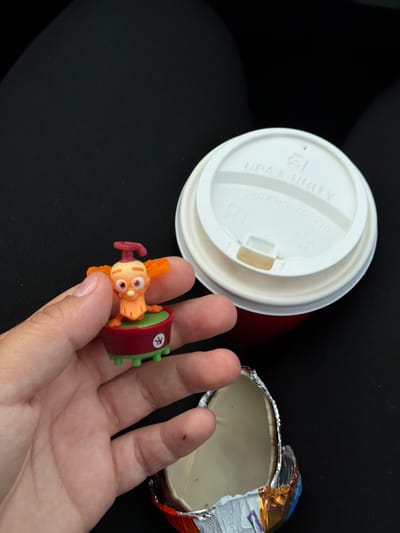 A person's hand holds a small, orange, cartoon-like toy figure with large eyes and orange wings. The figure is perched on a red and green platform with green legs. In the background, a white paper cup with a lid sits next to a red cup. Partially visible at the bottom of the image is the foil wrapper of an opened chocolate egg. The scene appears to be indoors, possibly in a vehicle, with a black, textured surface filling the background. The location in Усть-Сыны, Russia, is not discernable from the visual information provided.
