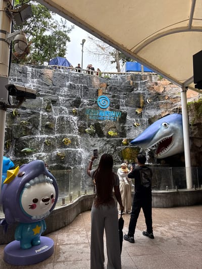 The image depicts an outdoor viewing area at the Qingdao Underwater World in Shinan District, Qingdao, China. The scene features a large, multi-tiered artificial rock-face waterfall with water cascading downwards. Embedded within the rock-face are various artificial marine creatures, including colorful fish and sea turtles. Centered on the rock-face is a blue circular logo with a spiral design, accompanied by the Chinese characters "青岛海底世界" and the English text "Underwater World." To the right, a large, realistic blue and white shark head sculpture with an open mouth and exposed teeth protrudes from the rock-face. To the left, a purple and light blue anthropomorphic character statue, resembling a creature in a shark costume with a yellow star, stands on a matching base.
In the foreground, three individuals are present. A woman with dark, braided hair, wearing a grey short-sleeved top and grey wide-leg pants, stands with her back to the camera, holding a smartphone vertically to capture the scene. A folded black umbrella hangs from her left hand. To her right, a man with dark hair, wearing a black t-shirt with a graphic design and black pants, also stands with his back to the camera, holding a smartphone horizontally to photograph the scene. Between these two, a third person, wearing light-colored clothing, holds a small child dressed in yellow, both facing the waterfall.
The viewing area is covered by a light-colored fabric canopy supported by metal poles, and the ground is paved with grey and beige square tiles. On the far left, two surveillance cameras are mounted on a vertical pole. Above the waterfall, on an elevated platform, several additional people are visible behind a railing, with trees and an overcast sky in the background.