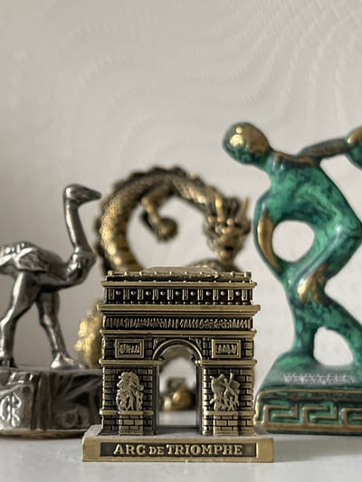 A close-up, eye-level shot displays a collection of miniature decorative objects against a blurred, white background. In the foreground, a small, metallic replica of the Arc de Triomphe stands prominently, with the inscription "ARC DE TRIOMPHE" visible on its base. To the left of the Arc, a metallic figurine of an ostrich with its head raised is partially visible. Behind the ostrich and the Arc, a golden, coiled dragon figurine is centered. To the right of the Arc, a green and gold statue of a discus thrower in motion is depicted. The objects are arranged on a white surface, and the lighting suggests an indoor setting, possibly on a shelf or table. The image does not provide any information about the city of Новогорелово, Russia.