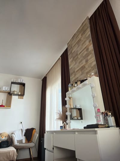 The image depicts an indoor scene, likely a bedroom or dressing area, with a focus on a vanity table and its surroundings. The room features white walls and a light-colored ceiling. On the left side of the frame, there are two floating shelves mounted on the wall. The upper shelf holds two white shopping bags with "PANDORA" branding. The lower shelf displays a small Eiffel Tower figurine, some red decorative items, and a small white container.

Dominating the right side of the image is a white vanity table with a large mirror that has integrated light bulbs around its perimeter. The surface of the vanity is cluttered with various cosmetic items, including bottles of makeup remover, brushes, perfumes, and other personal care products. A small black book or wallet is also placed on the vanity.

In the mid-ground, between the shelves and the vanity, is a light-colored chair with gold-colored legs. Several pieces of clothing, including a white top, a dark brown garment, and an orange item, are draped over the chair. A stuffed toy and a black bag are visible on a surface to the left of the chair, which appears to be part of a sofa or bench.

The background includes a tall window covered by two layers of curtains: a sheer white curtain closest to the window and a heavy brown curtain on the outer side. A section of the wall above the vanity is decorated with a textured panel made of horizontally stacked stone-like tiles. An electrical outlet is visible on the wall above the chair. The floor appears to be dark wood. The lighting suggests it is daytime, with natural light entering through the window.

There are no people visible in the image. The overall impression is of a well-organized personal space, perhaps a dressing area in a private residence. The specific location of Zmeitsa, Bulgaria, is not directly indicated by any visual elements within the image itself.