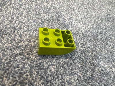 A bright green LEGO brick rests on a grey and white flecked carpet. The brick is oriented so that its top surface, with its signature circular studs, is facing upwards and slightly to the right. The brick has a boat-like shape with angled sides and a flat bottom. There are five visible studs on the top surface, arranged in a 2x2 pattern with one stud offset to the right. The lighting suggests it's daytime, casting a soft shadow from the brick onto the carpet. The carpet has a textured, plush appearance. There is no discernible activity or text in the image. The location context is Gostagayevskaya, Russia.