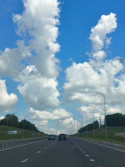 The image depicts a highway scene under a bright blue sky with scattered white clouds, presumably on the outskirts of the city of Шихазаны, Russia. The perspective is from a vehicle traveling on the road, showing a multi-lane highway with a central white line marking the lanes. Several cars are visible on the road ahead, including a dark SUV and a silver sedan. The roadside features a metal guardrail and a grassy embankment leading up to a line of trees and power lines in the distance. Streetlights line the highway at regular intervals.