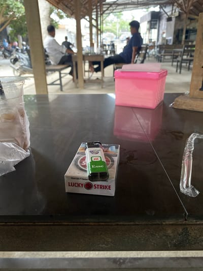 The image captures a casual indoor or semi-outdoor setting, likely a cafe or eatery, in Gampongaceh, Indonesia. The perspective is from a table, looking towards the back of the establishment.

In the immediate foreground, resting on a dark table surface, are several objects: a clear plastic cup containing a beverage and wrapped in plastic, a green and white cigarette lighter, and a pack of Lucky Strike cigarettes. A clear plastic straw or tube is also visible near the edge of the table. The cigarette pack prominently displays "LUCKY STRIKE," "Dibuat oleh PT Bentoel," and elements like "JESSE" and "Esse." The lighter also features the "Esse" brand logo.

Further back on the table, there is a pink plastic container. The background is blurred, showing several people and more tables and chairs. One individual in a blue shirt and dark pants is seen standing, while another in a white shirt is seated. The architecture includes wooden beams, suggesting an open or traditional structure. Beyond the seating area, hints of an outdoor space with parked motorbikes are visible. The scene portrays a daytime environment where people are gathered, possibly for dining or socializing, with smoking materials present on the table.