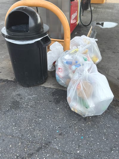 A black trash can with a domed lid sits next to several clear plastic trash bags filled with refuse. The trash can is positioned on a paved surface, and the trash bags are scattered to its right. In the background, a red and white gas pump with the "Pilot" logo is visible, along with a yellow safety barrier. A small puddle of liquid is present on the pavement near the gas pump. The scene appears to be an outdoor setting, likely at a gas station.