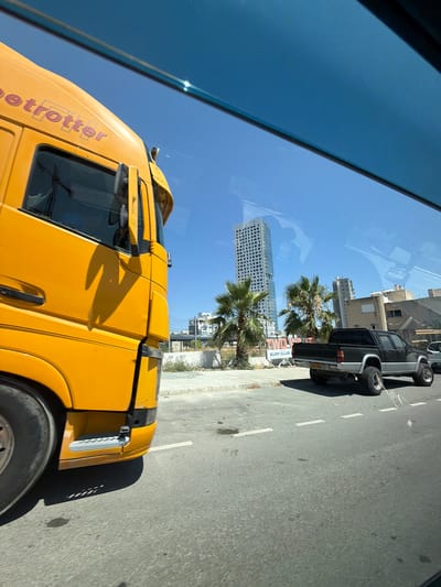 The image shows a bright yellow truck in the foreground, partially obscuring a street scene. The truck has the word "trotter" visible on its side in a dark pink font. A dark-colored pickup truck is visible on the road behind the yellow truck. In the background, a tall, modern high-rise building stands out against a clear blue sky, alongside other buildings and palm trees. The scene appears to be a city street in Mouttagiaka Community, Cyprus, during daytime. The road has white lane markings, and there are some shadows cast on the asphalt. The perspective suggests the photo was taken from inside another vehicle.