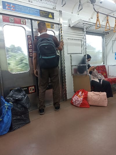 The scene is the interior of a train car in Bojong Gede, Indonesia. A man stands facing the doors, wearing brown pants and a blue backpack, holding the door open with his right hand. A woman wearing a hijab sits on a seat in the train car, with a red bag on the floor in front of her. There is a white handle on the top of the door and a black plastic trash bag on the floor next to the man. Another red bag with white lettering is on the floor next to the woman, and a pink bag is next to that. An open window is to the left of the man, providing a view of trees and greenery. The car is white with black lines. The woman's right hand is visible, holding something. The man is not looking at the camera. The woman is looking down at her hand.