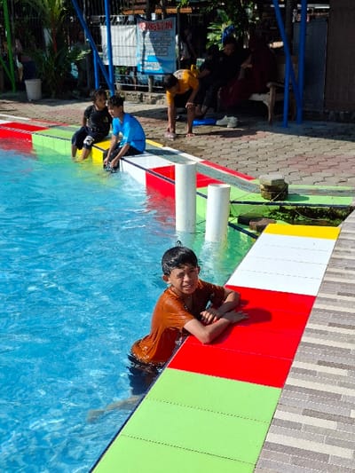 The scene shows a swimming pool in Kecamatan Tulung, Indonesia, located at latitude -7.592 and longitude 110.65. The pool is surrounded by a tiled deck, and is partially filled with water. A boy wearing a brown shirt is sitting on the edge of the pool with his arms resting on the tile. Three other boys are sitting on the edge of the pool, one wearing a blue shirt, one wearing a red shirt, and one wearing a white shirt. They are all looking at the camera. The pool appears to be made of concrete, and there are blue poles on the side of the pool. Behind the pool, there are green trees and a blue sky. The deck is made of concrete, and there are tiles of green, white, red, and yellow. The pool is empty except for the boy in the brown shirt. The scene appears to have been taken on a sunny day.