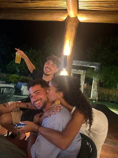 Three people are captured in an outdoor setting. One individual in a black shirt holds a drink, grinning with tongue slightly out. A woman with curly hair kisses the cheek of another man, who wears a blue and white checkered shirt, holds a phone, and smiles broadly. The woman is adorned with large, round earrings and a white dress. The background is filled with greenery and a stone structure. The photo is taken at night, illuminated by warm lighting.