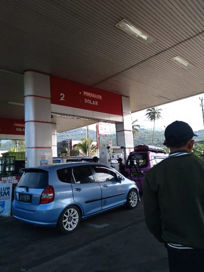 A blue car is at a gas station, next to a purple car. The gas station has a sign that says "PERTALITE SOLAR". The car has a license plate, and is near a trash can. A man in a green jacket and black hat stands next to the car. The background includes trees and a landscape. The image appears to be taken at a gas station near a scenic location.