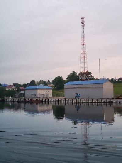 The image shows a waterfront scene in Ivanov Bor, Russia. A large, modern building with a blue roof and grey corrugated siding stands on a pier. To the left of this building is a smaller, lighter-colored building with a blue roof. In the background, several houses with red and blue roofs are visible amongst trees. Dominating the background is a tall, red and white lattice communication tower. The water in the foreground reflects the buildings and the tower. A small blue crane-like object is positioned on the pier in front of the large building. The sky is overcast and pale.