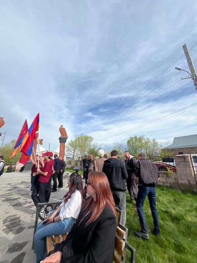 The image shows a group of people in what appears to be a memorial or park setting. Several people are seated on benches, while others stand nearby. Some individuals are holding Armenian flags. A set of khachkars (Armenian cross-stones) is visible in the background. The location is indicated to be in Akhuryan, Armenia. Overhead power lines are also in view.