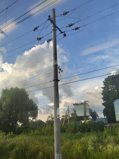 The image depicts a utility pole with multiple wires and insulators against a cloudy blue sky. The pole is positioned in the foreground and is gray and cylindrical. Below the pole, a lush green landscape of tall grass and various trees stretches towards the horizon. In the background, parts of buildings with blue roofs and smaller structures are visible amidst the trees, suggesting a rural or semi-rural setting. The scene appears to be located in the village of Konobeevo, Russia. There are no people or visible actions in the image. The lighting suggests it is daytime, possibly late afternoon, with the sun casting soft light through the clouds. Reflections are visible on the surface from which the image was taken, possibly a window.