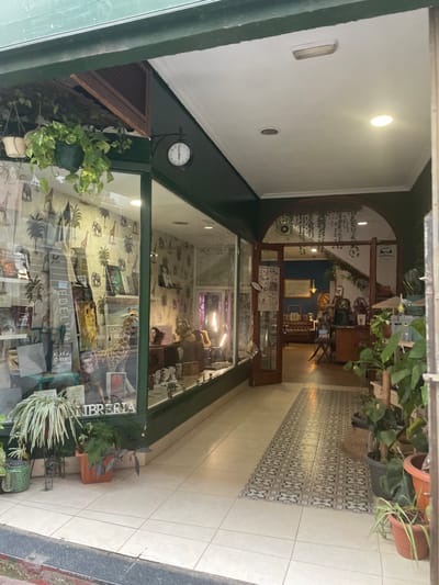 This image captures a view into a small storefront, likely a bookstore, from a covered outdoor or semi-enclosed walkway. The scene is set in what appears to be a pedestrian arcade or similar public space, possibly in Mérida, Spain, given the context.
The passageway leading to the shop has a clean, tiled floor. The central part features a long runner of white and grey patterned tiles, flanked by plain light-colored square tiles. The walls and ceiling of the walkway are white, with overhead light fixtures providing illumination.
The shop itself has a dark green facade and a large glass display window that offers a clear view of its interior. Visible through the glass, the shop is filled with books, arranged on shelves and in stacks. A prominent display within the window includes a tall giraffe statue, other decorative items, and potted plants. The word "LIBRERIA" (Spanish for 'bookstore') is prominently displayed in white lettering across the top of the shop's window. Other visible text includes "EDELVIVES" and "BLASCO IBANEZ" on book covers.
Inside, the walls are decorated with a pattern featuring tropical foliage like palm trees. An arched wooden doorway at the back leads to another section of the store, which also appears to house bookshelves, wooden furniture, and more plants, contributing to a natural and inviting atmosphere. The interior lighting appears to be artificial, with some fixtures casting a slight purple hue.
On the left side of the shop's exterior, a round, white-faced clock with black hands is affixed to the green frame, showing a time close to 12:40. Below the clock, a hanging potted plant with vibrant green leaves adds to the greenery. Potted plants are also visible outside the shop near the base of the window. The overall impression is that of a charming and thoughtfully decorated bookstore or gift shop.