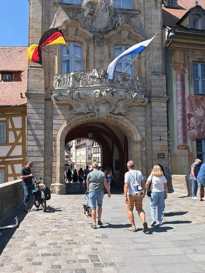 The image shows the historic Old Town Hall (Altes Rathaus) in Bamberg, Germany, which is situated on an island in the Regnitz River. The building is characterized by its ornate facade, featuring a large archway and balconies adorned with elaborate sculptures. Two flags are visible: a German flag and a blue and white flag, possibly representing Bavaria or the city of Bamberg.
In the foreground, several people are walking across a cobblestone bridge. A man in a dark t-shirt and blue jeans stands with a bicycle. Further ahead, a woman in a green t-shirt and denim shorts walks a small dog on a leash. Behind them, a couple walks arm-in-arm. The man wears a striped t-shirt and brown shorts, carrying a colorful backpack, while the woman wears a white t-shirt and blue pants. Other individuals are seen walking through the archway and in the distance. The environment is sunny with clear blue skies, and shadows are cast across the cobblestones. The architecture of the surrounding buildings, including half-timbered structures, is also visible.