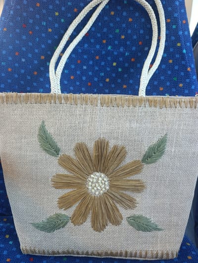 A close-up, eye-level, medium shot shows a handmade tan jute tote bag with a decorative straw flower applique and embroidered green leaves. The bag is placed on a blue fabric surface with a small, scattered pattern of red, yellow, and light blue dots. The tote bag has two braided white rope handles that extend upwards and out of the frame. The top edge of the bag is finished with a fringe of tan threads, and the bottom edge has a decorative stitch. The flower applique is centered on the bag, with a light-colored, textured center and light brown, straw-like petals. Two green embroidered leaves are positioned on either side of the flower. The background fabric appears to be a seat cushion or upholstery. The image provides no information about its location in Pinhal Novo, Portugal, or any people, actions, or interactions.