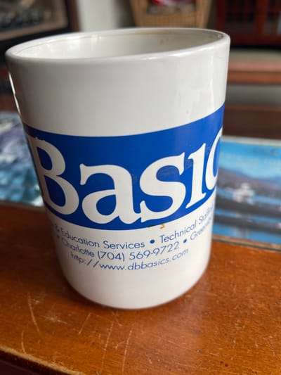 A white ceramic mug with a blue stripe across its middle sits on a wooden surface. The mug has text printed on it. The text reads "Basics" in large white letters on the blue stripe, and below that, smaller blue text says "& Education Services, Technical Staffing, Charlotte (704) 569-9722, Greensboro, http://www.dbbasics.com". The mug appears to be empty, and the wooden surface is a table. The background is blurred, showing what appears to be framed art and other objects on a shelf or in the background. The image is taken from a slightly elevated angle, looking down at the mug. There is no indication of the city of Brantingham, United States, in the image.