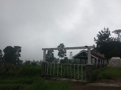 The scene depicts an outdoor environment in Joshi Vihir, India, under an overcast sky. In the immediate foreground, a dirt path or track is visible, leading to a metal gate. The gate features white or light-colored horizontal bars and is framed by two stone pillars with a horizontal wooden beam overhead, forming a simple entrance structure. To the left of the path and gate, dense green grass and various shrubs cover the ground.
Behind the gate and to the right, a single-story building is situated. This structure has a prominent gabled roof with a dark red or brown covering and appears to have light-colored walls. An open-sided shelter or porch area extends from the front of the building, supported by pillars, also with a gabled roof. To the far right of the building, a large, light-colored, irregular mound or rock formation is present on the grassy ground.
Numerous trees are scattered throughout the midground and background, exhibiting varying shades of green foliage. A distinct conifer or pine tree stands to the right of the building. The background shows a hazy, misty landscape, with hills or mountains densely covered in trees, fading into the atmospheric conditions. No people or animals are visible, and no discernible actions or interactions are occurring.