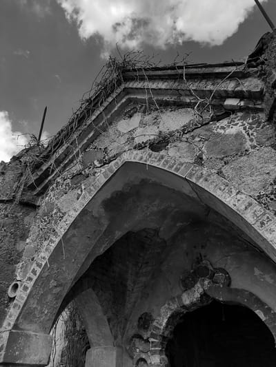A low-angle, black and white photograph depicts a decaying architectural structure against a backdrop of cloudy sky. The structure is heavily weathered, with crumbling stonework, brick elements, and exposed surfaces. Vegetation, including bare vines and branches, clings to the exterior. The structure includes an arched opening that leads into darkness. A small pipe protrudes from the lower right. The scene appears to be in a state of disrepair and abandonment. The location is Жалудоцкі сельскі Савет, Belarus.