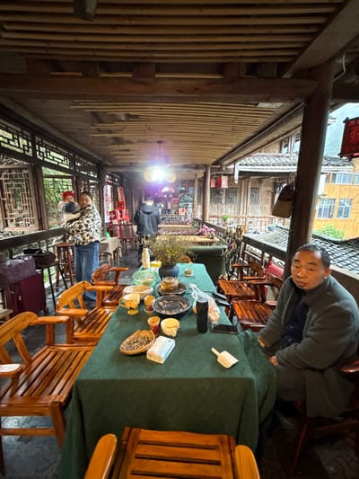This image depicts a covered outdoor dining area or balcony in Gongtan, China. The scene features three individuals and a table set with remnants of a meal, all within a traditional wooden architectural environment.

In the foreground, a long wooden table covered with a dark green tablecloth occupies the center and right. Scattered across the table are several used ceramic cups and bowls, a round metal grill grate, a dark teapot, a black thermos bottle, a plastic bag containing food, a woven basket with small dried items, a stack of packaged tissues, and a small white scoop-like object. To the right of the table, an adult male with short dark hair and a neutral expression sits facing left, wearing a dark green padded jacket over a blue shirt.

To the far left of the image, a woman stands holding a baby. The woman wears a patterned white and black jacket over blue jeans or a long skirt. The baby wears a red hat. Adjacent to them, a red suitcase and a brown bag rest on the floor. Further back in the mid-ground, another adult male stands with his back partially to the camera, wearing a dark jacket, facing into an illuminated interior area.

The environment consists of dark wooden beams and slats forming the ceiling overhead. Ornate wooden lattice screens and railings line the left side, with green foliage visible beyond. Traditional multi-story wooden buildings with tiled roofs are visible in the background, indicating the dense architecture of Gongtan. A bright light source, possibly a bare bulb, creates a glare in the middle-background. Red decorative banners with Chinese characters (including "2026") are visible on the left, near a smaller wooden table and chairs. Several wooden chairs with slatted backs and seats are placed around the main table and other smaller tables throughout the space.