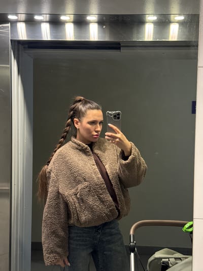 The image depicts a woman taking a mirror selfie in what appears to be an elevator, located in Gribovo, Russia. The woman, positioned slightly off-center to the left, is seen from the chest up, facing right and looking into the smartphone held by her right hand. Her dark hair is styled in a high ponytail that transitions into a long, thick braid extending down her left side. She wears a textured, light brown, collared fleece jacket over a darker brown or black inner layer, paired with blue denim jeans. A small earring is visible in her right ear. Her right hand, with manicured nails, holds a silver-colored smartphone with multiple camera lenses, angled to capture her reflection. In the reflected environment, overhead, a horizontal strip of four circular recessed lights illuminates the scene. The background behind the woman appears to be a plain, dark grey wall. To the lower right of the reflection, the brown handle and grey fabric of a baby stroller are visible, with a bright green accessory clipped to the handle. A partial blue rectangular object is reflected on the far right edge of the mirror. Metallic trim borders the reflective surface.