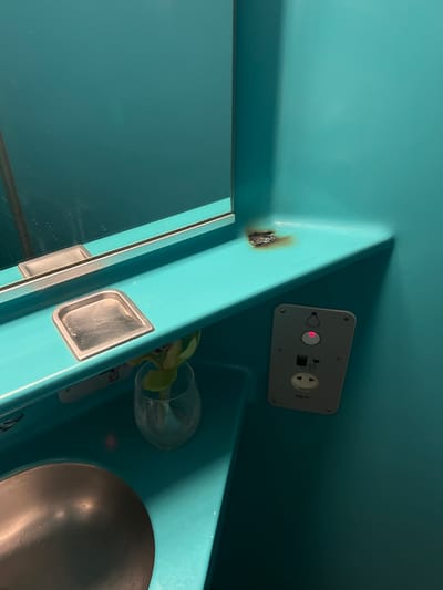 A teal-colored bathroom interior shot at an oblique angle. A gray outlet panel with a lightbulb icon, a red light, and a 230V outlet is mounted near a corroded area. A clear glass containing a green faux plant sits beside a metallic bowl. Below, two rectangular silver handles are visible on a teal surface, suggesting a cabinet or drawer. Lighting creates stark contrasts and shadows throughout.