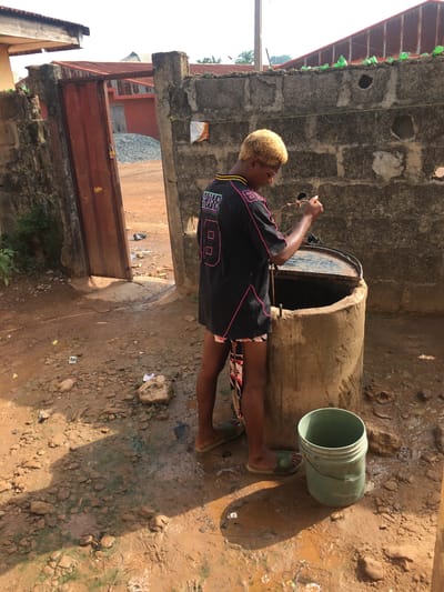 The image captures an outdoor scene in Isieke, Nigeria, illuminated by bright daylight, casting strong shadows. A person with short, bleached blonde hair and dark skin is featured, dressed in a black t-shirt or jersey with pink trim, stylized graphics, and the number "00," along with patterned shorts and green slide sandals.
They are positioned beside a large, cylindrical, concrete-like structure, likely a well or water storage drum, and appear to be drawing water using a rope that extends into its dark opening. A light green plastic bucket lies on the reddish-brown, unpaved dirt ground nearby. The ground is uneven, littered with rocks and debris, and shows damp, muddy areas from spilled water.
The background consists of rough concrete block walls, some bare and some painted. A corrugated metal roof is visible, adorned with green plastic bottles along its edge, possibly for water collection or decoration. A rustic red metal gate or door can also be seen in the distance. The overall setting suggests a utilitarian area within a residential or communal environment.