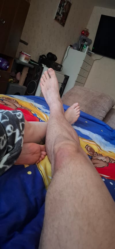 The image shows a close-up view of a person's legs crossed, lying on a bed with a colorful duvet. The person's feet are visible, with one foot resting on top of the other. The duvet has a pattern that appears to be cartoon characters. In the background, a dresser with drawers, a television, and some decorative items are visible, suggesting a bedroom setting. The overall lighting is dim, indicating it might be evening or nighttime. There are no specific landmarks or objects that would definitively place the scene in Марʼіна Горка, Belarus.