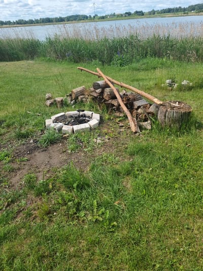 The image captures an outdoor scene near a body of water in Kalniškiai, Lithuania. In the foreground, a circular fire pit constructed from light-colored bricks is visible, containing ashes and remnants of burnt wood. To the right of the fire pit, a pile of chopped firewood is stacked, accompanied by a large tree stump and two long, rough-hewn wooden poles leaning against the woodpile. The ground is covered in green grass, with some patches of bare earth and scattered weeds. In the background, tall reeds and grasses grow along the edge of a calm body of water. Beyond the water, a distant treeline borders a gently rolling landscape under a cloudy sky. A wind turbine can be seen on a hill in the far distance. There are no people or animals present in the image.