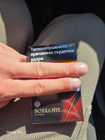 The image shows a light-skinned hand holding a black pack of Sobranie London cigarettes. The pack displays a Bulgarian health warning: "Тютюнопушенето причинява сърдечни удари" (Smoking causes heart attacks). The hand and cigarette pack are positioned over light-colored, textured fabric, consistent with woven trousers. Direct sunlight illuminates the scene, casting shadows from the hand and pack onto the fabric below. A "Galaxy S25" watermark is present in the lower-left corner. The specific location within Novo Delchevo, Bulgaria, is not discernible from the image.