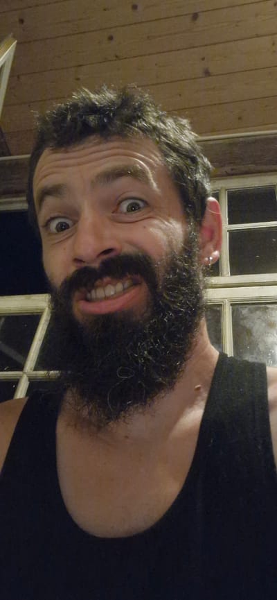 The image is a close-up, selfie-style photograph of a man with dark, messy hair and a full beard. He is wearing a black tank top. His eyes are wide open, and his mouth is open in a wide, toothy grin, suggesting a humorous or surprised expression. He has a small earring in his left ear. In the background, there is wooden paneling on the ceiling and a window with multiple panes. The lighting appears to be artificial and dim. The image does not contain any identifiable landmarks or features that would allow for specific location within Wattenwil, Switzerland.