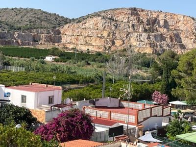 This media file depicts a sunny day in a rural landscape near Urbanització La Carrasquera, Spain. The scene is dominated by a large, multi-tiered quarry carved into a hillside, its rocky layers exposed and bathed in sunlight. Below the quarry, a flat expanse of lush green trees, likely an orchard, stretches across the middle ground. A small, white house with a red roof is visible within this green expanse.

In the foreground, several buildings with red-tiled roofs are visible, some with white walls and decorative patterned balustrades. Bright purple bougainvillea flowers cascade over one of these structures, adding a vibrant splash of color. Other elements in the foreground include trees, a streetlamp, and various awnings or canopies providing shade. In the distance, more greenery and some structures, possibly more homes or recreational areas, are partially visible.

The overall impression is one of a warm, clear day, with the bright sunlight casting sharp shadows. The presence of the quarry suggests a former or ongoing industrial activity, contrasting with the residential and agricultural elements. The blue sky indicates fair weather. No people are visible in the frame, and there is no discernible text.