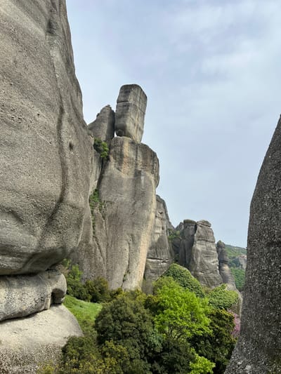 This image presents a dramatic natural landscape characterized by colossal, smooth grey rock formations under an overcast sky. The setting strongly suggests the unique geological features of Meteora in Kastraki, Greece.
The dominant subjects are the immense rock pillars and outcrops. On the left side, a remarkable formation resembles a towering stack of rounded, grey boulders, with the uppermost section appearing to balance on a narrower base, silhouetted against the sky. The rocks exhibit a weathered, somewhat granular texture with subtle horizontal striations in places.
Interspersed among the grey rock structures is an abundance of vibrant green vegetation. Lush trees and dense foliage, displaying various shades of green from bright lime to deep forest green, cling to the rock faces and grow in crevices, indicating fertile ground despite the harsh rock environment. A small cluster of purple or pink blossoms adds a delicate splash of color to the distant greenery on the right.
The sky is a uniform, soft light grey, indicating an overcast or cloudy day, which provides diffused lighting across the scene without harsh shadows. The foreground is dominated by the textured surface of another large, smooth grey rock, suggesting the photograph was taken from a high vantage point, looking out over the impressive geological formations.
No people or man-made objects are visible, emphasizing the untouched natural grandeur of the landscape. The scene conveys a sense of awe and the immense scale of these ancient rock formations. There is no visible text in the image.